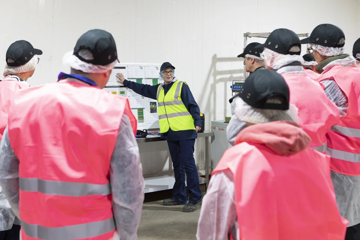 Middle aged woman presenting to investors in a factory setting, pointing to a white board filled with technical data.